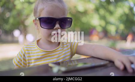 Petite fille dans les lunettes de soleil de mamans apprend la langue étrangère en répétant des mots à partir du téléphone portable. Portrait en gros plan d'une fille assise en ville Banque D'Images