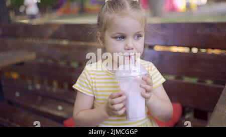 Une petite fille boit un milkshake à travers une paille. Portrait en gros plan d'une petite fille mignonne assise sur le banc du parc et buvant du milk-shake. Odessa, Ukraine Banque D'Images