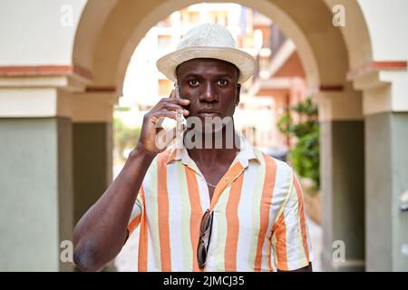 Un homme afro-américain parle au téléphone portable tout en se tenant dans la rue sous l'arche de la ville le jour de l'été Banque D'Images