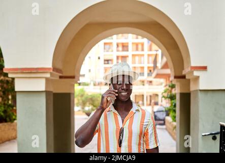 Un homme afro-américain parle au téléphone portable tout en se tenant dans la rue sous l'arche de la ville le jour de l'été Banque D'Images