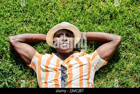 Par-dessus un homme afro-américain confiant en chapeau regardant vers le haut tout en se reposant sur une pelouse herbacée le jour d'été ensoleillé Banque D'Images