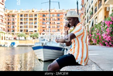 Vue latérale d'un homme d'affaires afro-américain souriant qui vérifie l'heure sur une montre-bracelet tout en ayant une conversation téléphonique sur le port près de la mer avec le bateau Banque D'Images