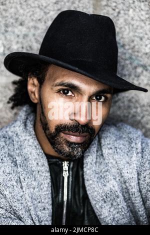 Homme hispanique à barbe sérieux, vêché de vêtements et de chapeaux élégants, regardant l'appareil photo contre un mur de pierre dans la rue Banque D'Images