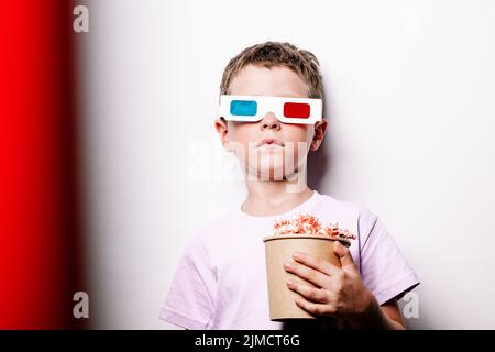 Enfant de 3D verres colorés mangeant du pop-corn savoureux dans un seau en carton tout en se tenant sur fond blanc dans un studio léger Banque D'Images