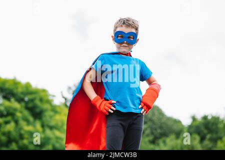 De dessous brave petit garçon avec masque et cape rouge volant main sur la taille contre le ciel gris le jour d'été dans le parc Banque D'Images