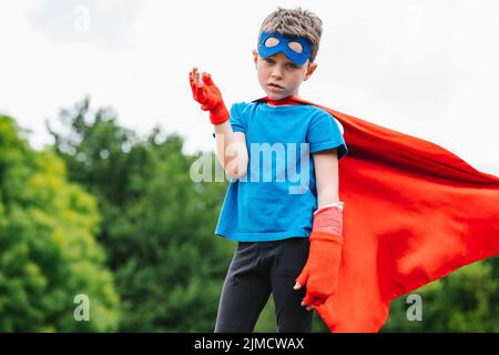 Petit super-héros avec cape et bras de levage de masque regardant la caméra contre les arbres verts et ciel nuageux le jour d'été dans le parc Banque D'Images