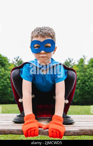 Petit super-héros avec cape et masque qui se croque contre des arbres verts et ciel nuageux le jour d'été dans le parc Banque D'Images