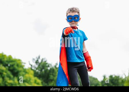 Petit super-héros avec cape et masque bras levant avec poing fermé regardant la caméra contre les arbres verts et ciel nuageux le jour d'été dans le parc Banque D'Images