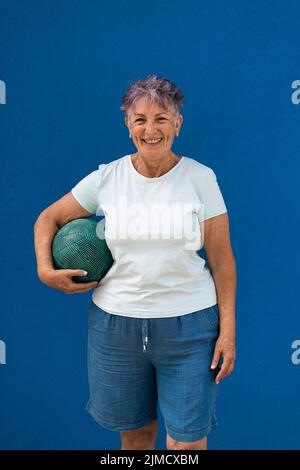 Femme sénior avec cheveux courts en short denim et t-shirt blanc debout sur fond bleu avec balle dans la main et sourire à l'appareil photo Banque D'Images