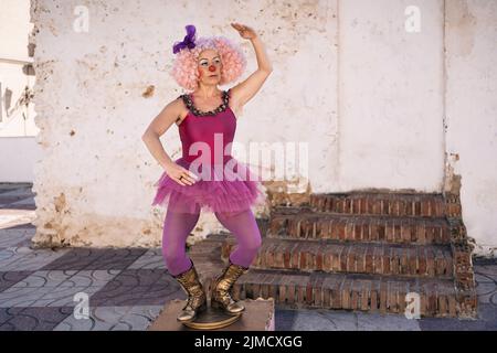 Corps entier d'artiste mime femelle mature en tutu et perruque rose avec maquillage brillant debout sur le piédestal avec bras relevé et vue loin Banque D'Images