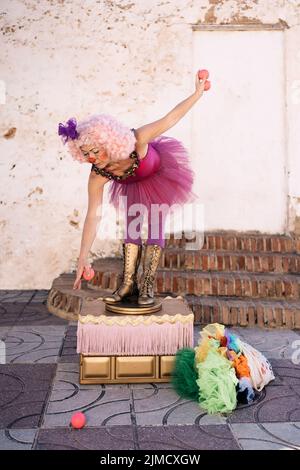 Ensemble complet de clown féminin d'âge moyen avec une tenue amusante et une perruque rose en forme de curly penchée vers l'avant tout en essayant de prendre le ballon pendant le spectacle de jonglage dans la rue Banque D'Images