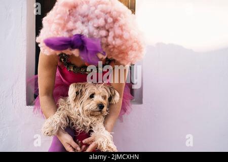Clown femelle adulte méconnaissable dans une perruque rose drôle et un costume qui embrasse un adorable chien moelleux tout en étant assis dans la rue après le spectacle Banque D'Images