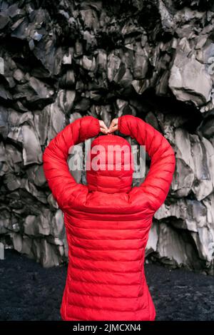 Vue arrière d'une femme voyageur méconnaissable en veste à capuchon rouge aux bras levés tout en admirant la falaise de basalte sur la plage de Reynisfjara en Islande Banque D'Images