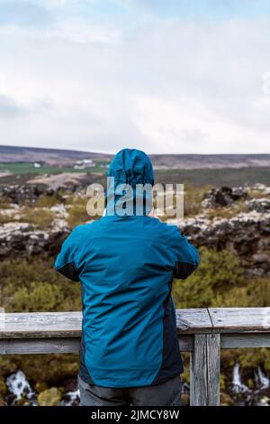 Vue arrière d'un voyageur masculin méconnaissable en veste à capuche, debout sur un point de vue en bois et admirant le paysage pittoresque du terrain volcanique vallonné avec W Banque D'Images