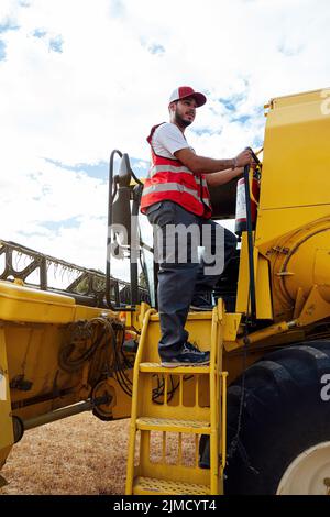 Depuis le dessous du côté de la carrosserie entière de l'opérateur de la moissonneuse-batteuse mâle pensive, debout de manière uniforme sur l'échelle de la machine professionnelle pour la collecte des grains dans l'agriculture Banque D'Images