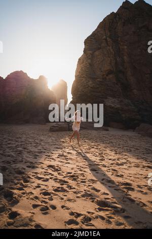Femme voyageur courant sur une côte sablonneuse avec des falaises rocheuses rugueuses près de la mer à l'heure du coucher du soleil le soir d'été ensoleillé Banque D'Images