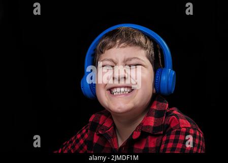 Un jeune garçon joyeux, joyeux et souriant dans une chemise à motif écossais qui écoute de la musique dans un casque circum-aural sans fil bleu sur un vêtement isolé Banque D'Images