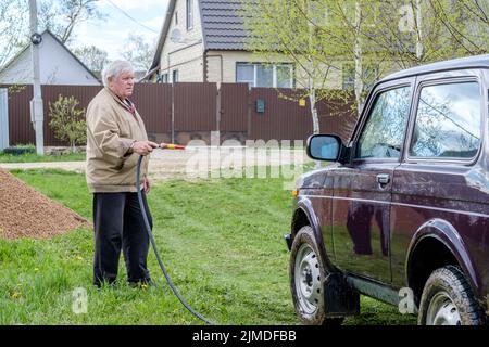 Un homme âgé aux cheveux gris lave une voiture à l'extérieur avec un tuyau d'arrosage. Banque D'Images