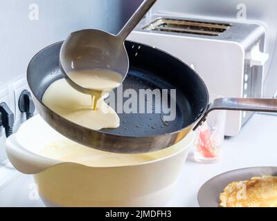 Faire couler une pâte d'un seau en métal sur une poêle graissée rouge-chaude. Le processus de cuisson des crêpes. Banque D'Images