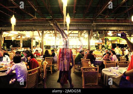 Un artiste portant une tenue traditionnelle Dayak passe devant les visiteurs assis autour de tables à manger, après un spectacle de danse traditionnelle du groupe sous-ethnique Murut appelé Anggalang & Magunatip à Kampung Nelayan, un restaurant de fruits de mer à Kota Kinabalu, en Malaisie. Banque D'Images