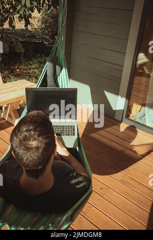 Un homme indépendant se trouve dans hamac et travaillant près de maison de campagne. Travail à distance Banque D'Images