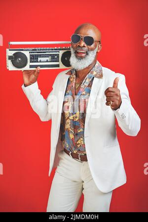 La musique est ce qui élève l'esprit. Photo studio d'un homme âgé portant des vêtements vintage en posant avec une boombox sur un fond rouge. Banque D'Images