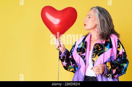 Un coeur qui aime est toujours jeune. Une femme âgée confiante et élégante tenant un ballon en forme de coeur sur un fond jaune. Banque D'Images