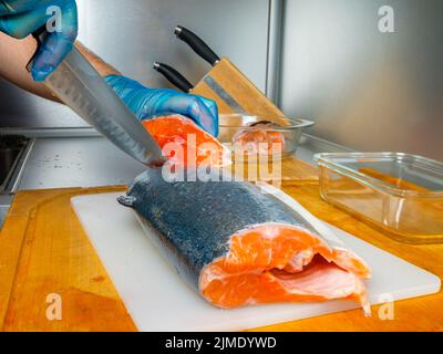 Les mains du cuisinier en gants coupent le corps du saumon avec un couteau. Banque D'Images