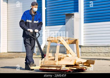 Un homme en vêtements de travail et un masque médical transporte une charge sur un chariot sur le territoire d'une base industrielle, le jour de l'été Banque D'Images