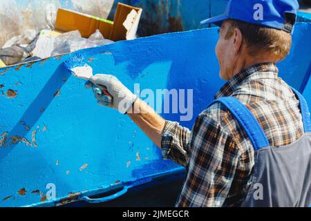 Un vieil homme dans une combinaison de travail peint une poubelle ou un conteneur dans la rue avec un pinceau. Banque D'Images