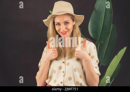 Femme blonde blanche portant un chapeau de soleil et une chemise estivale à pois debout à côté de la grande plante souriant à l'appareil photo montrant les pouces. Prise de vue en studio à arrière-plan sombre. Photo de haute qualité Banque D'Images