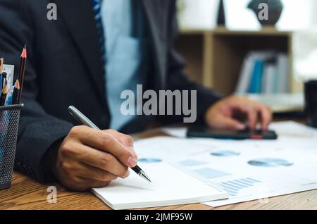 Homme d'affaires tenant un stylo écrivant un livre et un document graphique comptabilité finance papier analyse de travail graphique marketing investissement et rapport économie sur le bureau Banque D'Images
