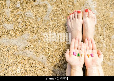 Les mains et les pieds des femmes avec une pédicure rouge sur fond de sable de mer et de surf. Dans les paumes des lettres avec les ins Banque D'Images