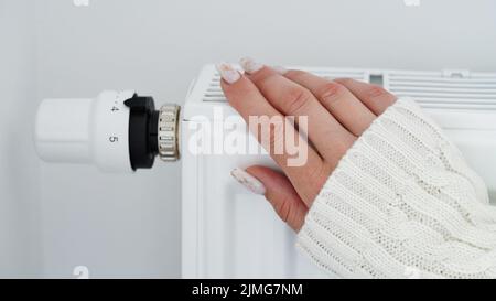 Gros plan d'une femme en chandail blanc chauffe ses mains sur la batterie sur fond de mur blanc.Macro photo des mains de la femme, copier l'espace.concept de s. Banque D'Images
