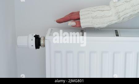 Gros plan d'une femme en chandail blanc chauffe ses mains sur la batterie sur fond de mur blanc.Macro photo des mains de la femme, copier l'espace.concept de s. Banque D'Images