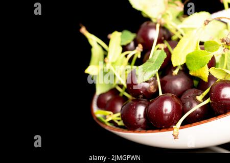 Plusieurs cerises douces mûres dans une plaque en céramique blanche, en gros plan, isolées sur un fond noir. Banque D'Images