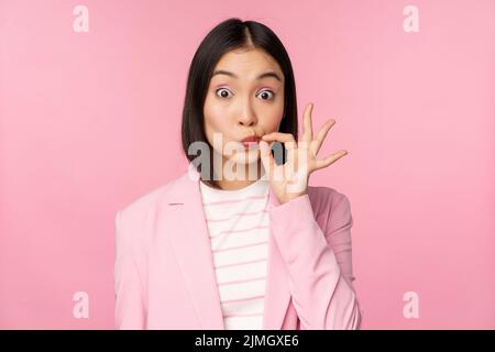 Portrait d'une femme d'entreprise asiatique montrant le sceau de la bouche, fermer les lèvres sur le geste clé, promesse garder secret, debout sur l'ba rose Banque D'Images