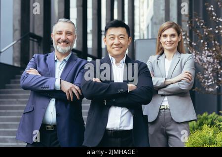 Portrait d'un patron asiatique réussi et heureux avec son équipe de rêve diversifiée, des gens d'affaires à l'extérieur du bâtiment de bureau debout avec les bras croisés et regardant l'appareil photo Banque D'Images