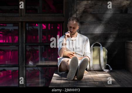 Le jeune homme est assis sur des escaliers en bois à l'extérieur, écrivant des notes dans un bloc de papier aux rayons du soleil Banque D'Images