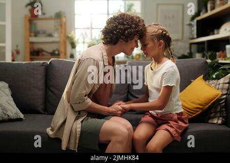 Jeune femme attentive en vêtements décontractés se pliant légèrement vers l'avant à sa jolie fille jeune assise devant et touchant son front Banque D'Images