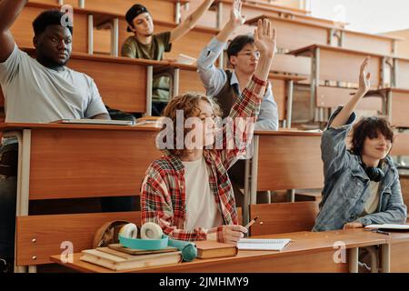 Plusieurs jeunes étudiants interraciaux levant la main pour poser des questions à l'enseignant sur le nouveau sujet tout en étant assis dans les rangs lors de la conférence Banque D'Images