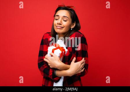 Jolie positive mignonne jeune femme brune isolée sur fond rouge mur portant blanc décontracté t-shirt et rouge et chemise noire Banque D'Images