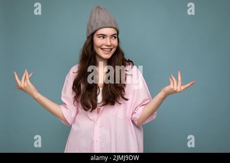 Portrait de belle positive gaie mignon sourire jeune femme brune dans une chemise élégante et chapeau gris hipster isolé sur bleu Banque D'Images