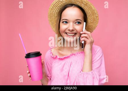 Portrait de belle positive émotionnelle jeune femme blonde portant un petit chemisier rose et chapeau de paille parlant sur smartphone pendant que d Banque D'Images