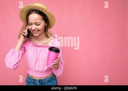 Photo de belle positive émotionnelle jeune femme blonde portant un chemisier rose et chapeau de paille parlant sur le téléphone cellulaire pendant la boisson Banque D'Images