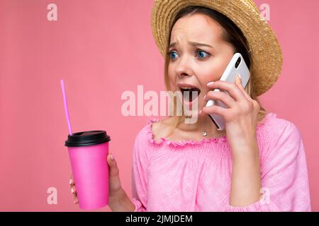 Portrait d'attrayant choqué émotionnel jeune blonde femme portant un petit chemisier rose et chapeau de paille parlant sur smartphone Banque D'Images