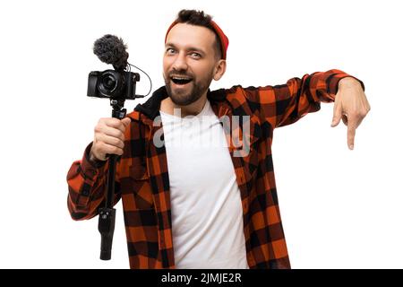 Homme d'âge moyen barbu blogueur avec une caméra et un microphone dans ses mains gesturant vers le bas sur un fond blanc Banque D'Images