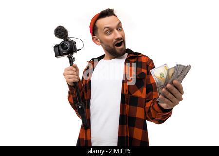 Jeune homme blogueur avec une caméra vidéo avec un microphone et de l'argent dans ses mains sur un fond blanc Banque D'Images