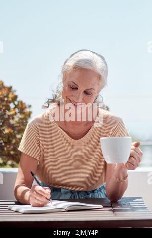 Vivez le genre de vie les meilleures ventes sont faites. Une femme mûre ayant le café et l'écriture dans un carnet sur le balcon à la maison. Banque D'Images