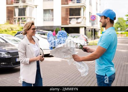 Un jeune liveur en uniforme bleu livre des fleurs à un client de femme Banque D'Images